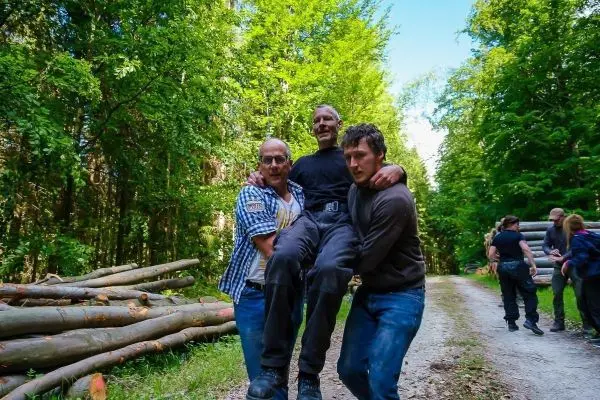 Drei Personen tragen einen Mann auf einem Waldweg zwischen Holzstämmen